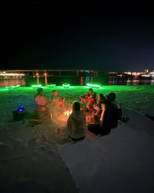Guests gathered around a campfire on the island at night, with glowing kayaks lined up along the shore