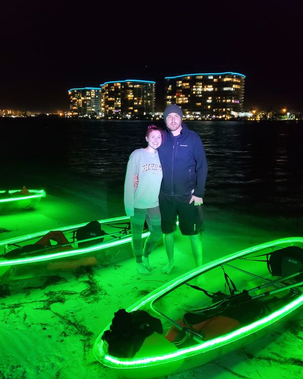 A smiling couple standing on the island beside their glowing kayaks, with the lit condos across the water