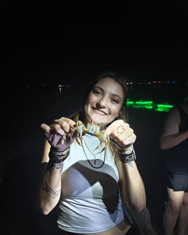 A smiling guest holding up a crab with both hands on the beach at night