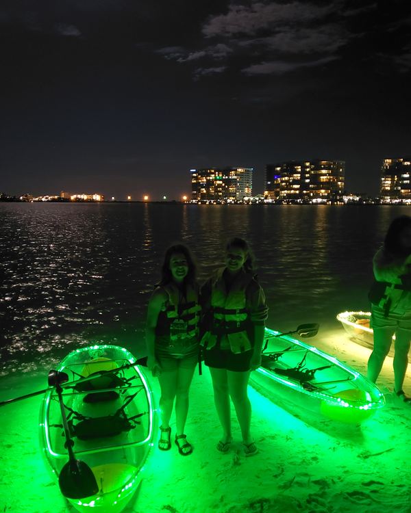 Two guests in life jackets standing on the island between glowing kayaks, with the skyline across the bay