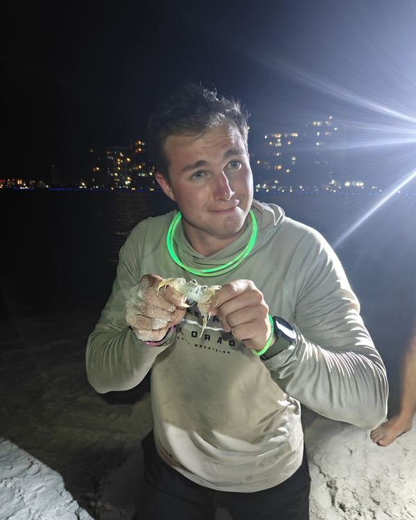 A grinning guest with a glow necklace holding up a crab he caught on the beach at night