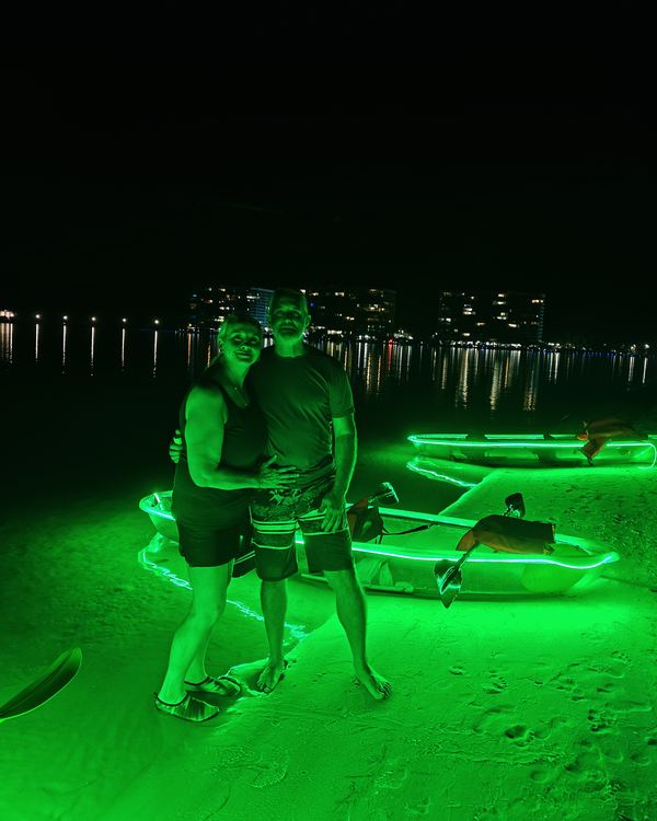 A couple standing arm in arm on the island next to glowing kayaks, city lights behind them